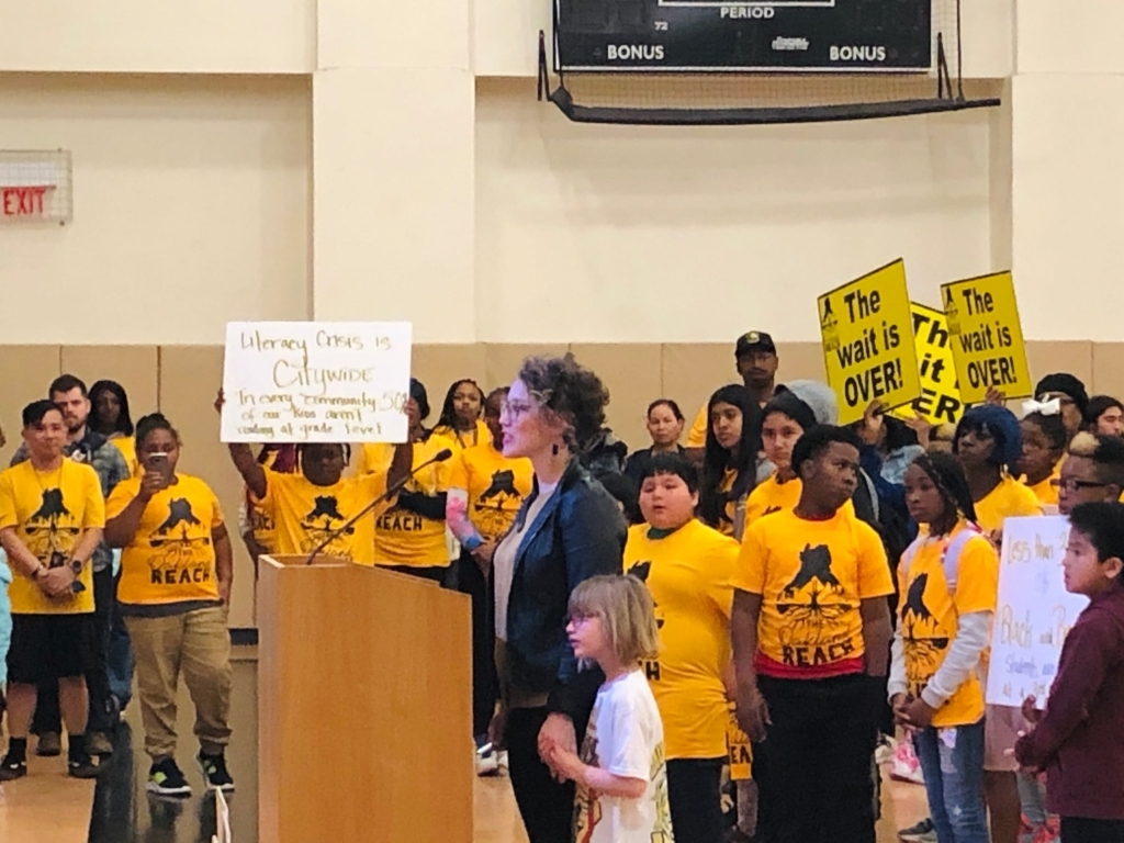 Cassie Perham Standing At A Podium Speaking In A Crowded Auditorium