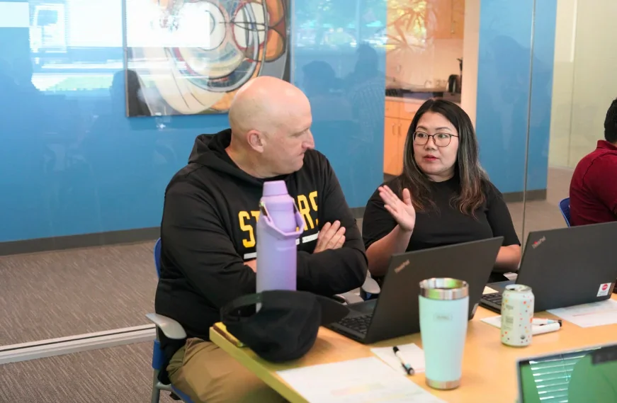 Principal Cheng Saechao speaks with Math Teacher Leader Alan Gates as they sit at a table behind their laptop computers, collaborating during a Math Community of Practice session at Highland Elementary.