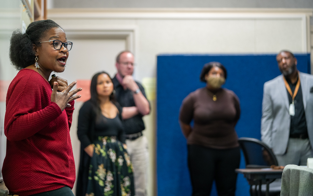 A black woman in a red top speaks to a group of four school principals in the background against a white wall with a blue panel.