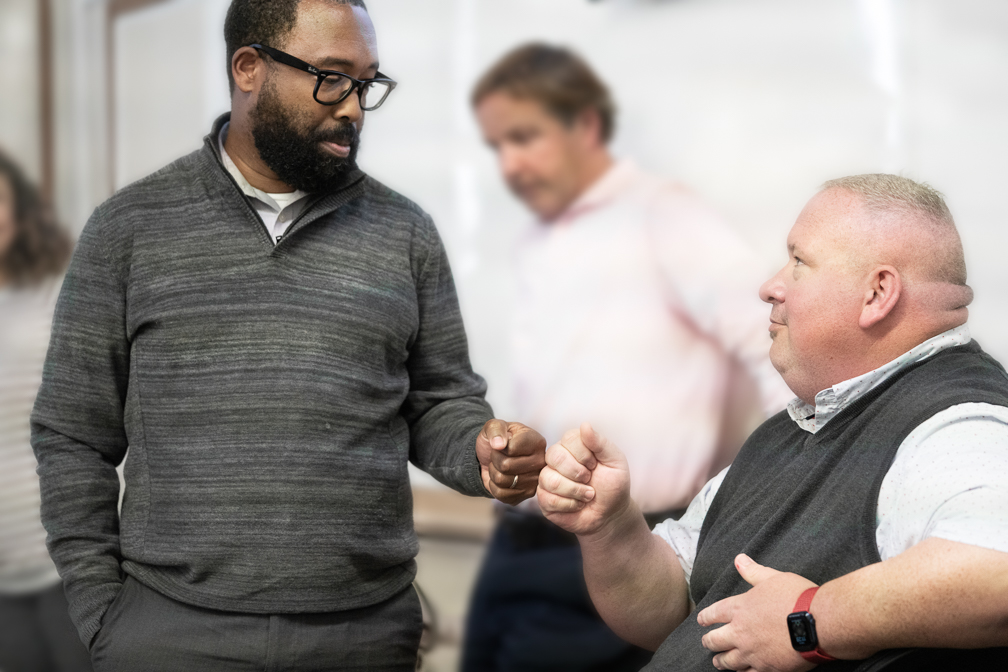 A bearded Black man wearing in a grey sweater fist bumps a white man in a grey sweater vest inside a classroom. A bearded Black man wearing in a grey sweater fist bumps a white man in a grey sweater vest inside a classroom.