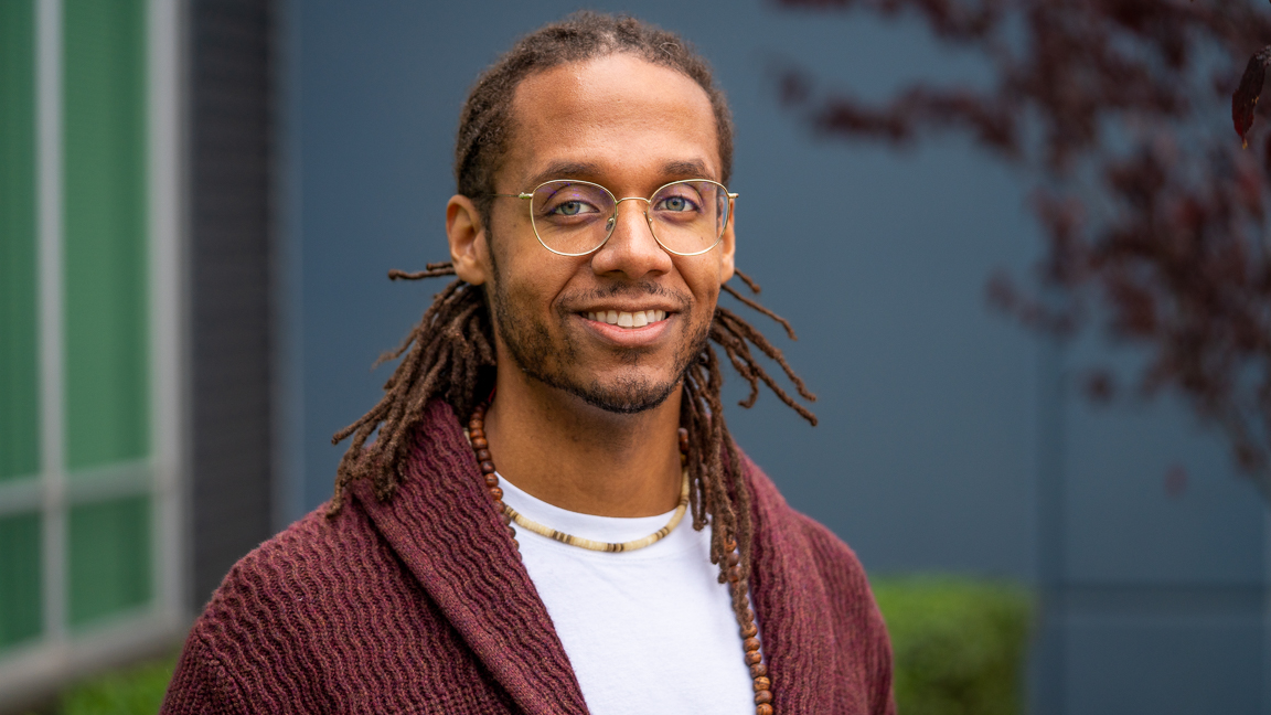 A Black man with dreadlocks wearing wire-rimmed glasse, a burgundy cardigan and beaded necklasses smiles in an outdoor setting in front of a dark blue wall edged with burgundy tree leaves and green shrubs.