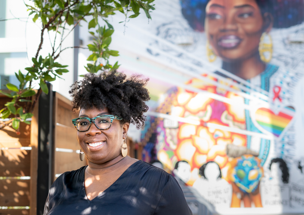 Una sonriente mujer negra de pelo rizado, con blusa negra de cuello de pico, gafas verdes con dibujos y grandes pendientes de latón, frente a un colorido mural que representa a una mujer negra en la pared de un patio al aire libre.