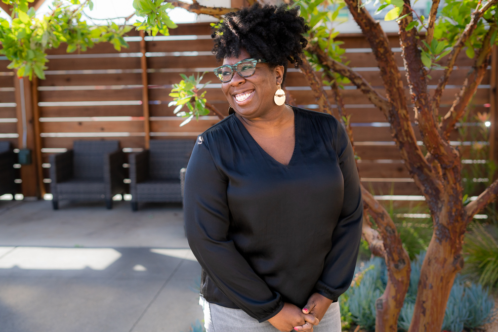 A grinning Black woman with curly hair wearing a black v-neck blouse, grey jeans, green patterned glasses, and large brass earrings stands in front of a tree and wooden fence on an outdoor patio.
