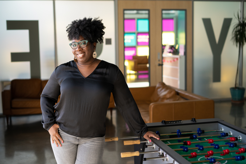 A smiling Black woman with curly hair wearing a black v-neck blouse, grey jeans, green patterned glasses, and large brass earrings stands near a foozball table in a spacious room in front of couches and a door to the RYSE Commons building.