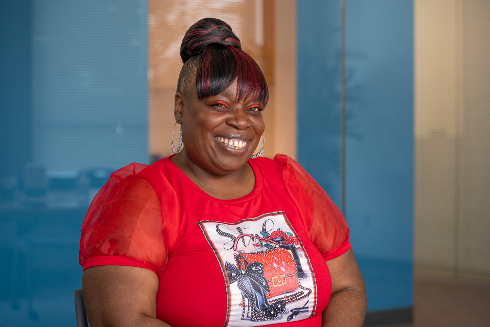 Una mujer negra sonriente lleva una blusa roja con una decoración estampada, sombra de ojos roja, pendientes de aro plateados y el pelo recogido en un moño con mechas rojas. El fondo es una pared azul borrosa con una puerta amarilla detrás de ella.
