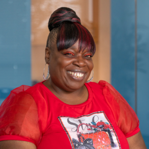 A smiling Black woman wearing a red blouse with a printed decoration, red eye shadow, silver hoop earrings, and her hair in a bun with red highlights. The background is a blurry blue wall with a yellow doorway behind her.