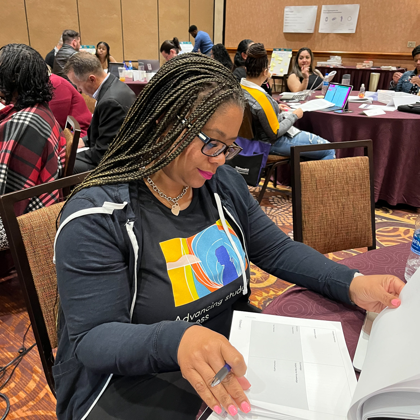Una mujer negra con largas trenzas y gafas está sentada en la mesa de una sala de conferencias revisando material impreso sobre planes de estudios e instrucción. Detrás de ella, en mesas cercanas, otras personas hacen lo mismo.