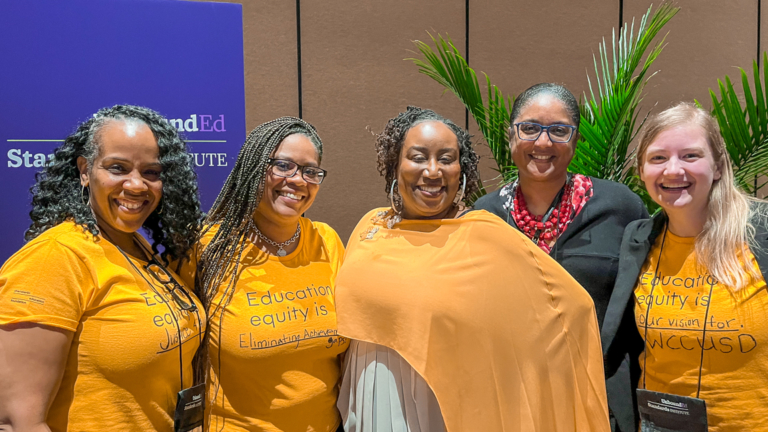 Cuatro mujeres negras y una blanca posan sonrientes en una sala de conferencias. Tres llevan camisetas doradas que dicen "La equidad educativa es" con una frase autocompletada escrita después.