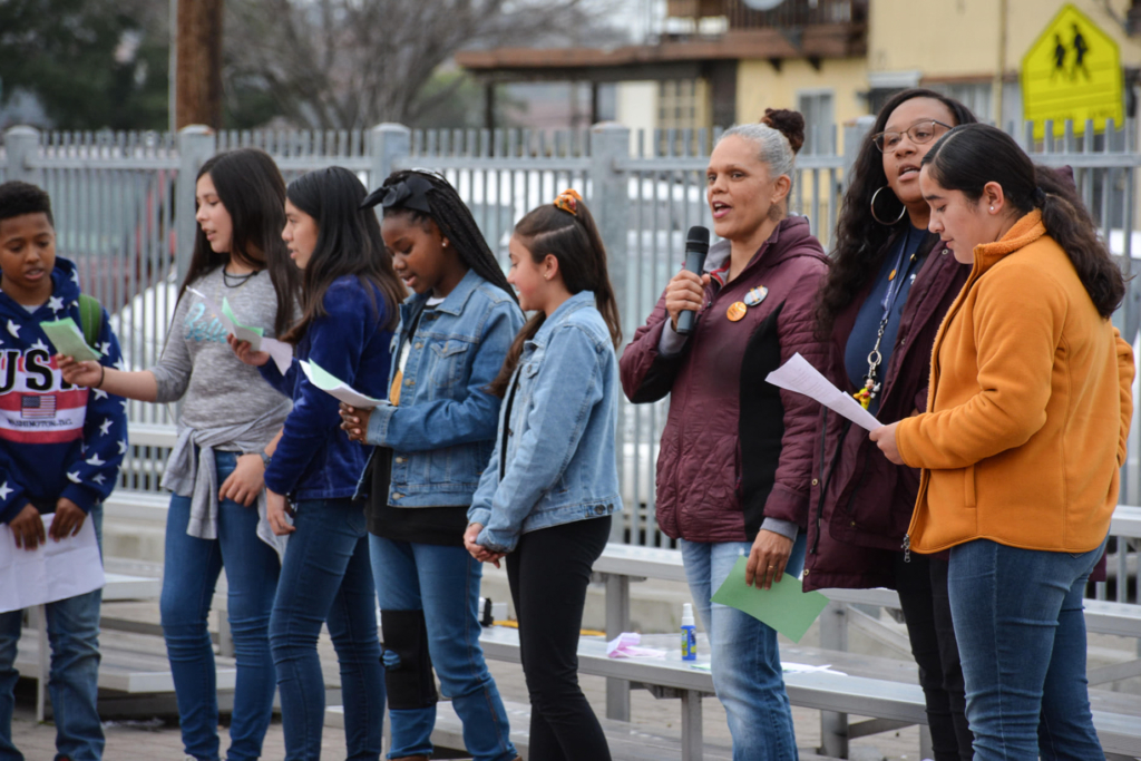 Tana Monteiro is standing outdoors in a burgundy jacket and blue jeans among a group of students. Monteiro is speaking into a hand-held microphone while the students read from paper cards.