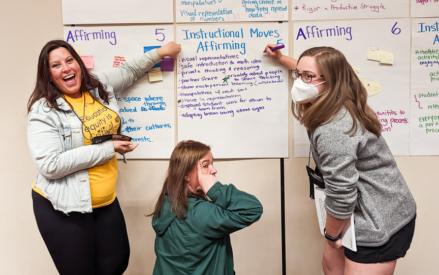 Tres mujeres sonrientes (una de ellas enmascarada) están delante de una pared señalando un póster con ideas y prácticas sobre planes de estudios culturalmente afirmativos.