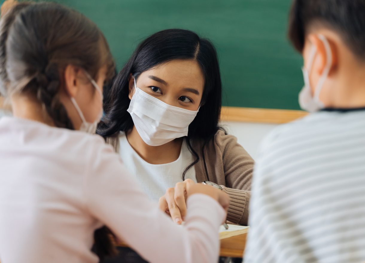 Close-up of Asian female teacher wearing face mask in school building tutoring primary student children.