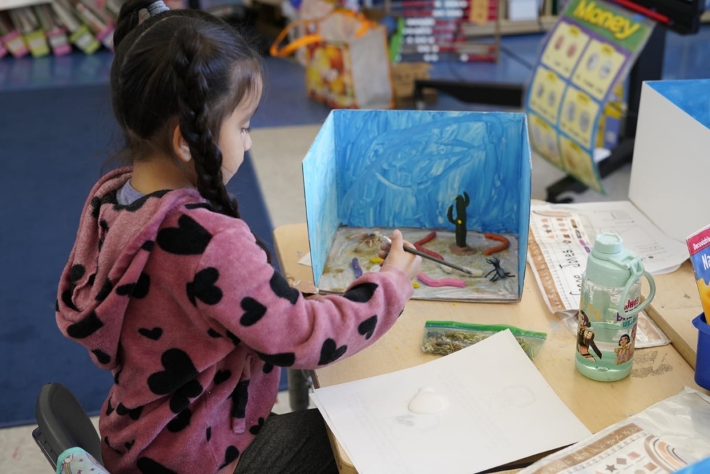 Student Leans Over A Desk And A Diorama Project